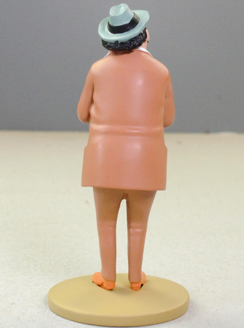 May include: A figurine of a person viewed from behind, wearing a light blue fedora with a black band, a tan suit jacket, and matching tan trousers. The figure stands on a circular tan base, with orange shoes visible at the bottom of the legs.