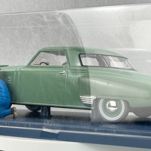 Pu&ograve; includere: Un modello di auto d'epoca verde con pneumatici a fascia bianca, esposto in una teca trasparente. Due figure in miniatura con tute blu e berretti sembrano lavorare sull'auto. Il modello &egrave; etichettato "1/18 BURAGO".