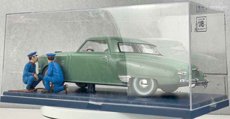 Pu&ograve; includere: Un modello di auto d'epoca verde con pneumatici a fascia bianca, esposto in una teca trasparente. Due figure in miniatura con tute blu e berretti sembrano lavorare sull'auto. Il modello &egrave; etichettato "1/18 BURAGO".