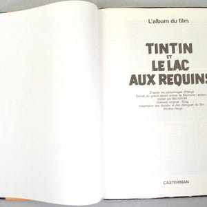 May include: Open book with the title page of a Tintin comic book. The title reads "TINTIN ET LE LAC AUX REQUINS" in large, bold letters. The book is open to a double-page spread with text and illustrations.