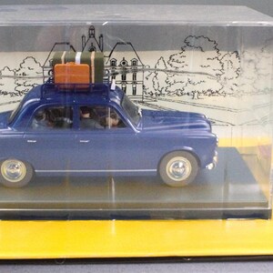 May include: A blue die-cast model car with luggage on the roof, displayed in a clear plastic case. The model sits on a yellow base with French text and a cartoon character. The car is a vintage style.