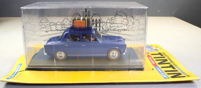 May include: A blue die-cast model car with luggage on the roof, displayed in a clear plastic case. The model sits on a yellow base with French text and a cartoon character. The car is a vintage style.
