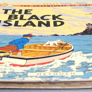 K&ouml;nnte beinhalten: Ein Vintage-Exemplar von "Die Schwarze Insel" von Herg&eacute;, aus der Reihe "Tim und Struppi". Das Cover zeigt ein Boot mit einer Figur und einem Hund auf dem Wasser, mit einer Burg auf einer Insel im Hintergrund. Das Buch hat einen roten R&uuml;cken und den Namen des Verlags.