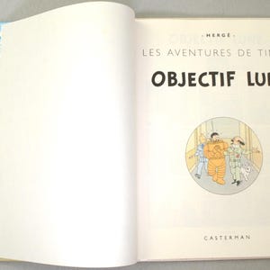 K&ouml;nnte beinhalten: Aufgeschlagenes Buch mit dem Titel "OBJECTIF LUNE" in gro&szlig;en schwarzen Buchstaben. Das Buch zeigt eine Comic-Illustration von drei Figuren in einem Kreis. Der Einband hat blau-wei&szlig; gestreifte R&auml;nder mit gerahmten Illustrationen.