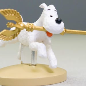 Puede incluir: Figura de perro blanco con nariz negra y boca abierta, sosteniendo un bast&oacute;n dorado con alas. El perro est&aacute; montado sobre una base beige, dando la impresi&oacute;n de vuelo. El bast&oacute;n tiene un extremo decorativo.