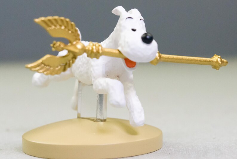 Puede incluir: Figura de perro blanco con nariz negra y boca abierta, sosteniendo un bast&oacute;n dorado con alas. El perro est&aacute; montado sobre una base beige, dando la impresi&oacute;n de vuelo. El bast&oacute;n tiene un extremo decorativo.