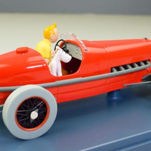 Pu&ograve; includere: Un'auto da corsa rossa vintage con un pilota in camicia gialla e una figura di cane bianco. L'auto ha ruote grigie con raggi neri e finiture argentate. Il modello &egrave; su una base blu.