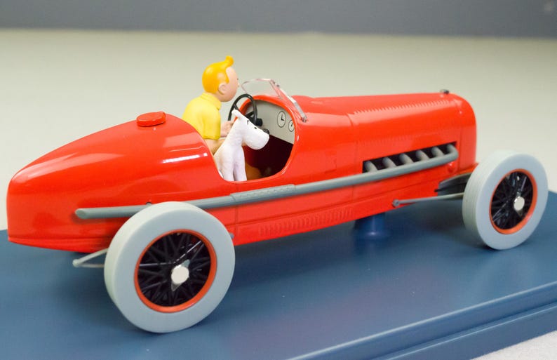 Pu&ograve; includere: Un'auto da corsa rossa vintage con un pilota in camicia gialla e una figura di cane bianco. L'auto ha ruote grigie con raggi neri e finiture argentate. Il modello &egrave; su una base blu.
