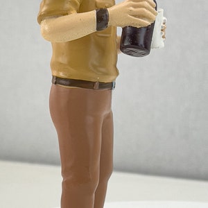 May include: A figurine of a man wearing a white and yellow captain's hat and a brown shirt and pants. He is holding a bottle of dark liquid and a white cup.