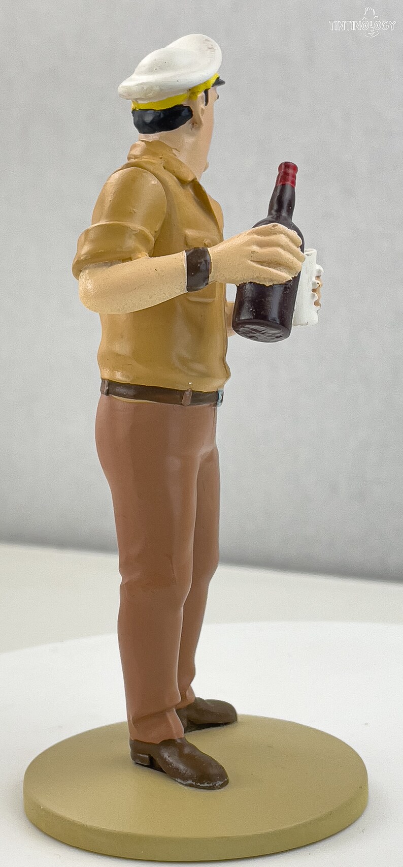 May include: A figurine of a man wearing a white and yellow captain's hat and a brown shirt and pants. He is holding a bottle of dark liquid and a white cup.