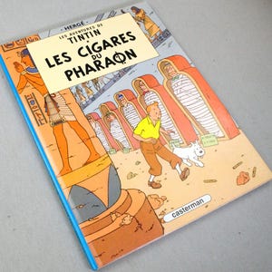 Pode incluir: Banda desenhada "Les Cigares du Pharaon" de Herg&eacute;. A capa apresenta uma ilustra&ccedil;&atilde;o de Tintin, Milu e cenas eg&iacute;pcias em laranja, amarelo e azul. Publicado por Casterman.