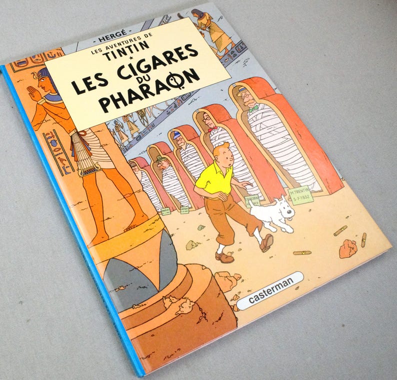 Pode incluir: Banda desenhada "Les Cigares du Pharaon" de Herg&eacute;. A capa apresenta uma ilustra&ccedil;&atilde;o de Tintin, Milu e cenas eg&iacute;pcias em laranja, amarelo e azul. Publicado por Casterman.