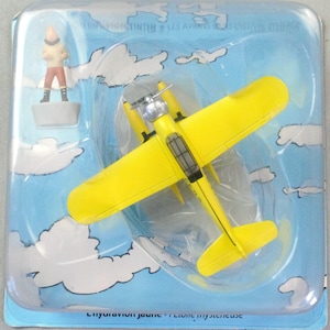 Op de afbeelding: Een felgeel speelgoedvliegtuig met zwarte details en een zilveren propeller, verpakt in een blauwe plastic verpakking met een stripfiguur. De verpakking heeft een lucht- en wolkenontwerp, met tekst aan de boven- en onderkant.