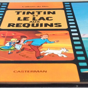 May include: A vintage hardcover book titled "Tintin et le Lac aux Requins" in French. The cover features a colorful illustration of Tintin, Snowy, and other characters running. The book has a black spine and a yellow border.