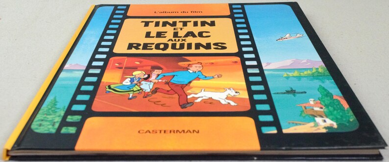 May include: A vintage hardcover book titled "Tintin et le Lac aux Requins" in French. The cover features a colorful illustration of Tintin, Snowy, and other characters running. The book has a black spine and a yellow border.