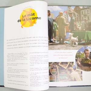 May include: An open book with a blue and white cover. The interior pages feature illustrations and text in French. One page has the title "Le m&acirc;t de la Licorne" in yellow and black. The other page shows a market scene with people and a dog.