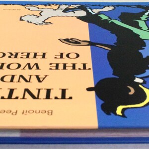 Puede incluir: Un libro de tapa dura titulado "Tint&iacute;n y el Mundo de Herg&eacute;" con una cubierta azul y una ilustraci&oacute;n de dibujos animados de Tint&iacute;n. El lomo del libro es azul y las p&aacute;ginas son blancas. La cubierta tiene una secci&oacute;n naranja claro.