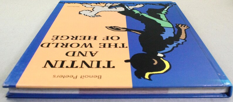 Puede incluir: Un libro de tapa dura titulado "Tint&iacute;n y el Mundo de Herg&eacute;" con una cubierta azul y una ilustraci&oacute;n de dibujos animados de Tint&iacute;n. El lomo del libro es azul y las p&aacute;ginas son blancas. La cubierta tiene una secci&oacute;n naranja claro.