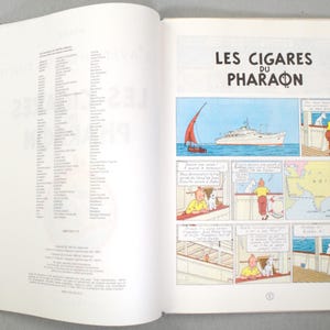 Pode incluir: Livro aberto com uma hist&oacute;ria em quadrinhos intitulada "Les Cigares du Pharaon". A hist&oacute;ria em quadrinhos mostra um barco no mar, um mapa da &Aacute;sia e personagens de desenhos animados. A p&aacute;gina esquerda tem uma lista de texto.