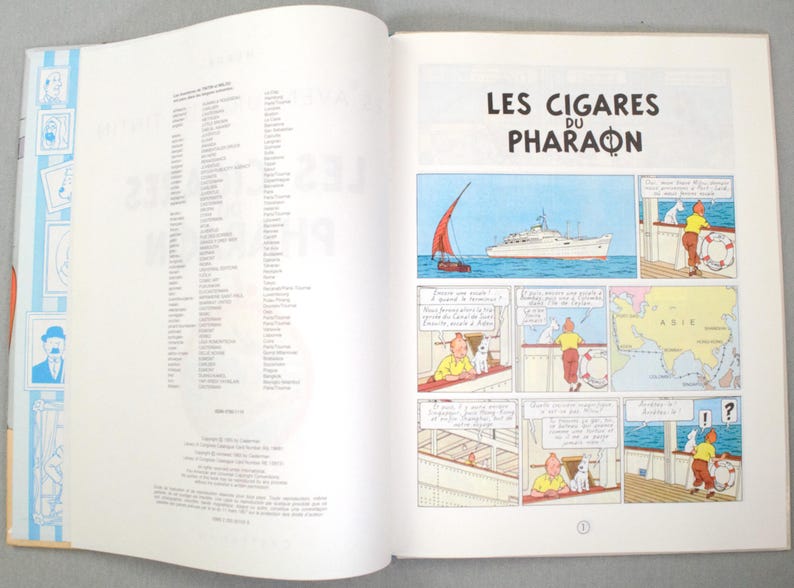 Pode incluir: Livro aberto com uma hist&oacute;ria em quadrinhos intitulada "Les Cigares du Pharaon". A hist&oacute;ria em quadrinhos mostra um barco no mar, um mapa da &Aacute;sia e personagens de desenhos animados. A p&aacute;gina esquerda tem uma lista de texto.