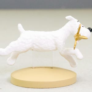 Puede incluir: Figura blanca de un perro corriendo, con una h&eacute;lice dorada en la boca. El perro tiene una nariz negra y est&aacute; montado sobre una base circular beige. Es un objeto decorativo.