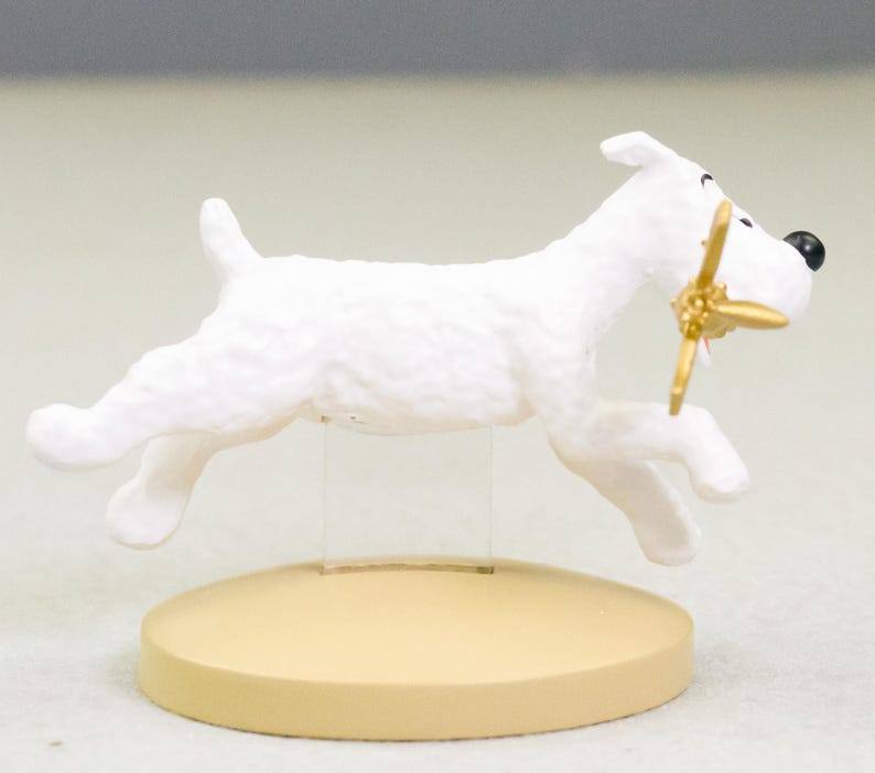 Puede incluir: Figura blanca de un perro corriendo, con una h&eacute;lice dorada en la boca. El perro tiene una nariz negra y est&aacute; montado sobre una base circular beige. Es un objeto decorativo.