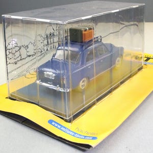 May include: A blue die-cast model car with luggage on the roof, displayed in a clear plastic case. The car is on a yellow and black cardboard base. The case has a drawing of a building on the side.