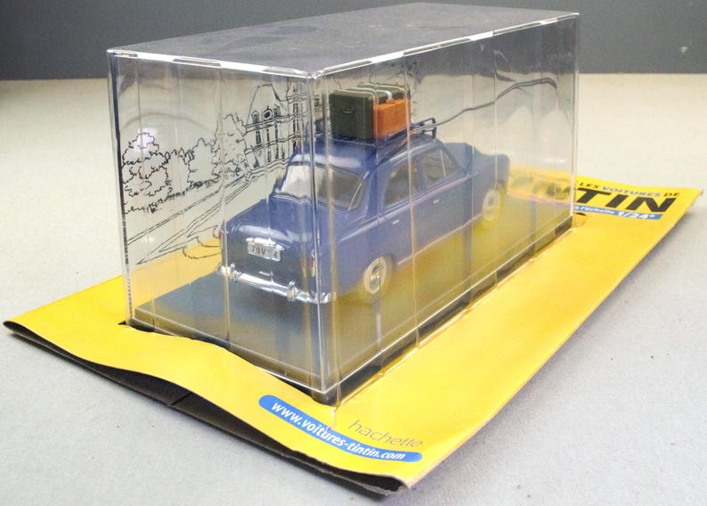 May include: A blue die-cast model car with luggage on the roof, displayed in a clear plastic case. The car is on a yellow and black cardboard base. The case has a drawing of a building on the side.