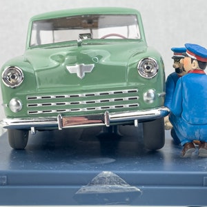 Pu&ograve; includere: Modello di auto verde in stile vintage con dettagli cromati e un emblema argentato. Due figurine in uniforme blu e berretto sembrano ispezionare l'auto. Su una base blu.
