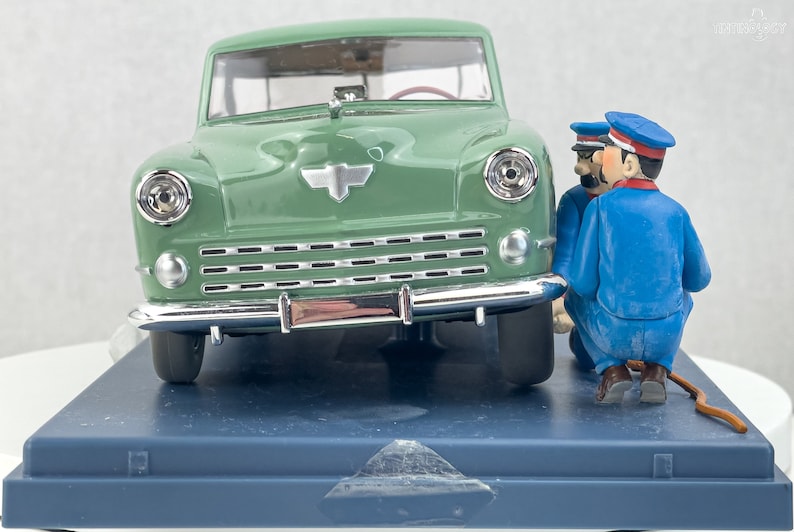 Pu&ograve; includere: Modello di auto verde in stile vintage con dettagli cromati e un emblema argentato. Due figurine in uniforme blu e berretto sembrano ispezionare l'auto. Su una base blu.