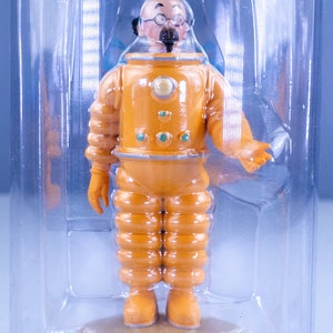 May include: A yellow astronaut figurine with a black mustache and glasses, standing on a brown base inside a clear plastic container.