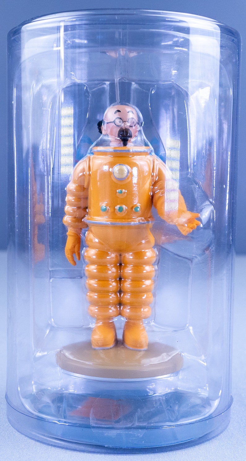 May include: A yellow astronaut figurine with a black mustache and glasses, standing on a brown base inside a clear plastic container.
