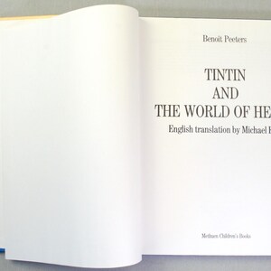 Puede incluir: Un libro abierto con el t&iacute;tulo "TINTIN AND THE WORLD OF HERG&Eacute;" en negro. El nombre del autor, Benoit Peeters, y del traductor, Michael Farr, tambi&eacute;n est&aacute;n presentes. El libro tiene una encuadernaci&oacute;n azul.