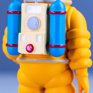 May include: A yellow astronaut figurine with a large, round head and a backpack with two blue oxygen tanks. The backpack has a cream-colored panel with a red button and a circular window.