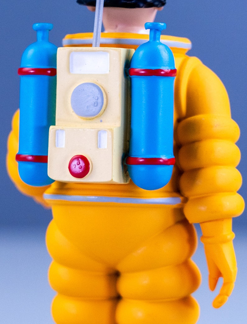 May include: A yellow astronaut figurine with a large, round head and a backpack with two blue oxygen tanks. The backpack has a cream-colored panel with a red button and a circular window.
