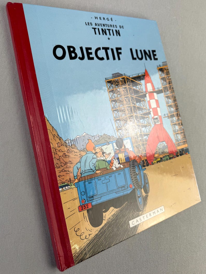 May include: A hardcover book titled "OBJECTIF LUNE" with a red spine. The cover features a blue jeep with characters, a red and white rocket, and the text "HERGE - LES AVENTURES DE TINTIN". The book is published by Casterman.