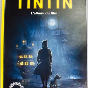May include: A book cover with the title "LES AVENTURES DE TINTIN" in yellow. The image shows a person and a dog standing before a ship at night. The text "L'album du film" is also visible on the cover.