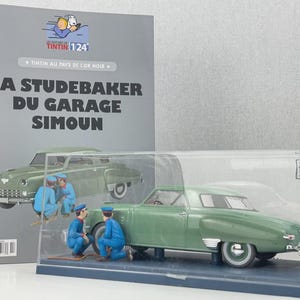 Pu&ograve; includere: Un modello di auto Studebaker verde in una vetrina trasparente, con tre figure in miniatura in tuta blu. Sullo sfondo un libro con il titolo "LA STUDEBAKER DU GARAGE SIMOUN", con un disegno dell'auto.