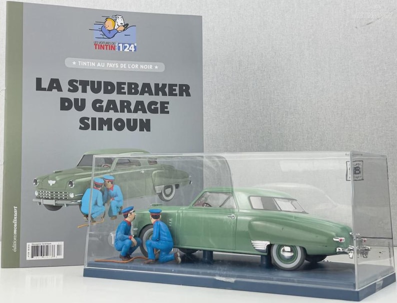 Pu&ograve; includere: Un modello di auto Studebaker verde in una vetrina trasparente, con tre figure in miniatura in tuta blu. Sullo sfondo un libro con il titolo "LA STUDEBAKER DU GARAGE SIMOUN", con un disegno dell'auto.