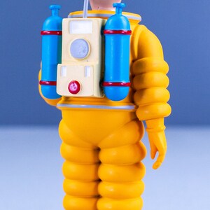 May include: A yellow astronaut figurine with a black hairpiece, a blue and red oxygen tank, and a white antenna. The figurine is standing on a tan base.