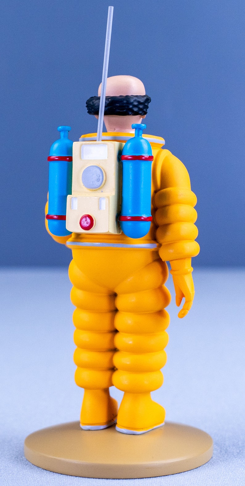 May include: A yellow astronaut figurine with a black hairpiece, a blue and red oxygen tank, and a white antenna. The figurine is standing on a tan base.