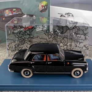 Puede incluir: Modelo de coche negro vintage con neum&aacute;ticos blancos, expuesto sobre una base azul. El modelo es una r&eacute;plica a escala 1:24, con detalles interiores visibles. Una vitrina transparente y una caja con una ilustraci&oacute;n de coche est&aacute;n en el fondo.