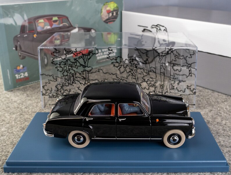 Puede incluir: Modelo de coche negro vintage con neum&aacute;ticos blancos, expuesto sobre una base azul. El modelo es una r&eacute;plica a escala 1:24, con detalles interiores visibles. Una vitrina transparente y una caja con una ilustraci&oacute;n de coche est&aacute;n en el fondo.