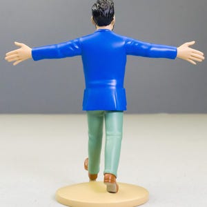 以下が含まれることがあります： 後ろ姿の男性のフィギュア。腕を広げています。鮮やかな青色のジャケット、薄緑色のズボン、茶色の靴を履いています。フィギュアは、ニュートラルな背景に対して、ベージュ色の円形の台座の上に立っています。