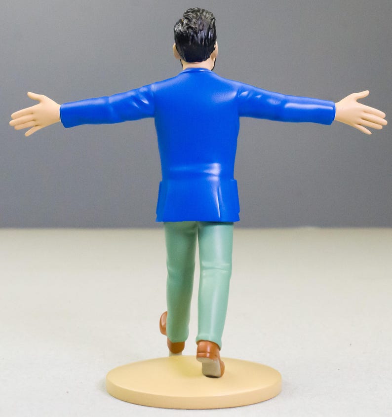 以下が含まれることがあります： 後ろ姿の男性のフィギュア。腕を広げています。鮮やかな青色のジャケット、薄緑色のズボン、茶色の靴を履いています。フィギュアは、ニュートラルな背景に対して、ベージュ色の円形の台座の上に立っています。