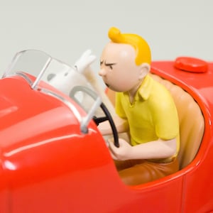 Pu&ograve; includere: Un'auto da corsa giocattolo rossa con parabrezza trasparente. Una statuetta di una persona con i capelli gialli e una camicia gialla guida l'auto. Un cane bianco &egrave; sul sedile del passeggero. L'auto ha un sedile rosso e un volante nero.