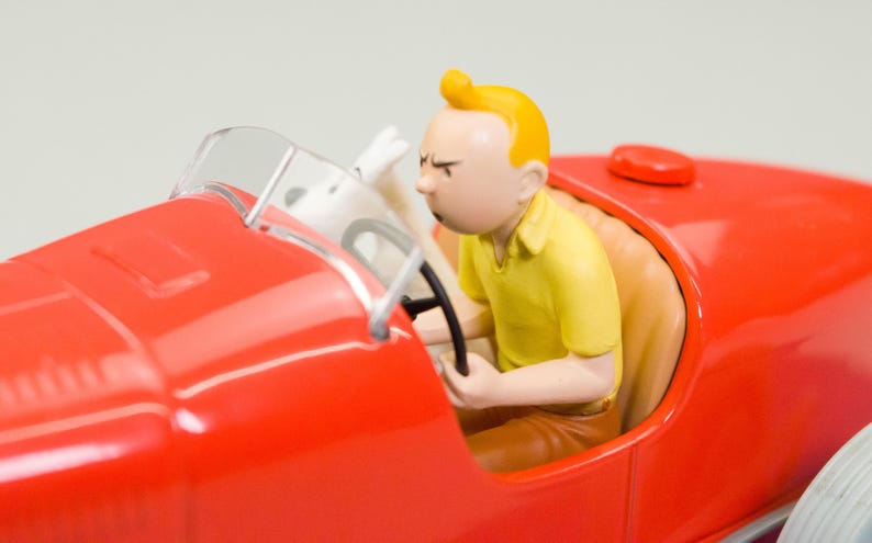 Pu&ograve; includere: Un'auto da corsa giocattolo rossa con parabrezza trasparente. Una statuetta di una persona con i capelli gialli e una camicia gialla guida l'auto. Un cane bianco &egrave; sul sedile del passeggero. L'auto ha un sedile rosso e un volante nero.