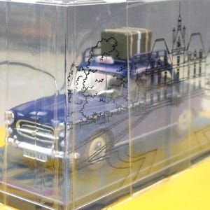 May include: A blue vintage car model with a luggage piece on top, encased in a clear plastic box. The box features a black line drawing of a building and trees. The model sits on a yellow base.