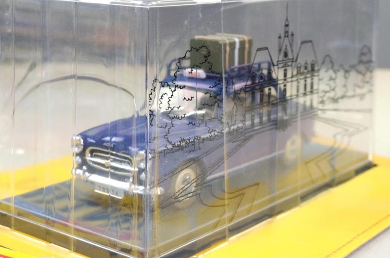 May include: A blue vintage car model with a luggage piece on top, encased in a clear plastic box. The box features a black line drawing of a building and trees. The model sits on a yellow base.