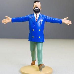 以下が含まれることがあります： 黒いひげと髪の男性のフィギュア。青いダブルブレストのジャケット、赤と白のストライプのネクタイが付いた白いシャツ、緑のズボンを着用しています。フィギュアは、腕を広げて、タン色のベースの上に立っています。